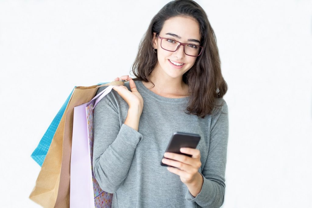 woman wearing glasses, carrying shopping bags in one hand and holding a smartphone in the other