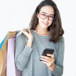 woman wearing glasses, carrying shopping bags in one hand and holding a smartphone in the other