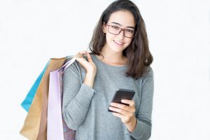 woman wearing glasses, carrying shopping bags in one hand and holding a smartphone in the other