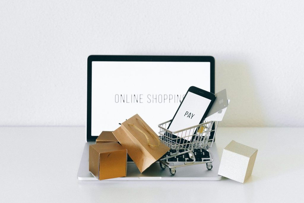 miniature boxes and a small shopping cart holding a smartphone with the word "pay" on the screen, with a laptop in the background displaying "online shopping"
